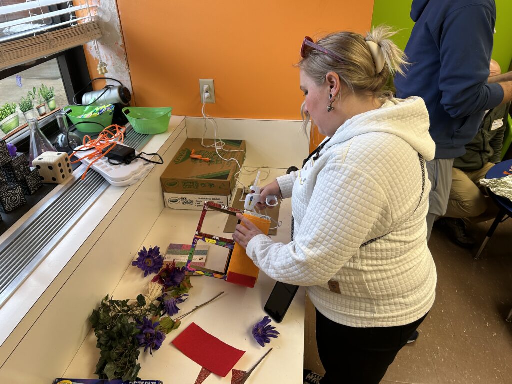 A woman stands at a workbench with crafting materials.