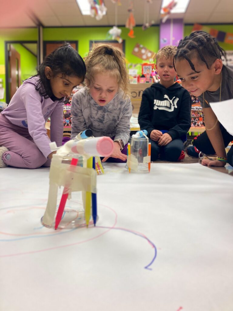 Four elementary school students kneel on a large sheet of white paper and play with craft materials