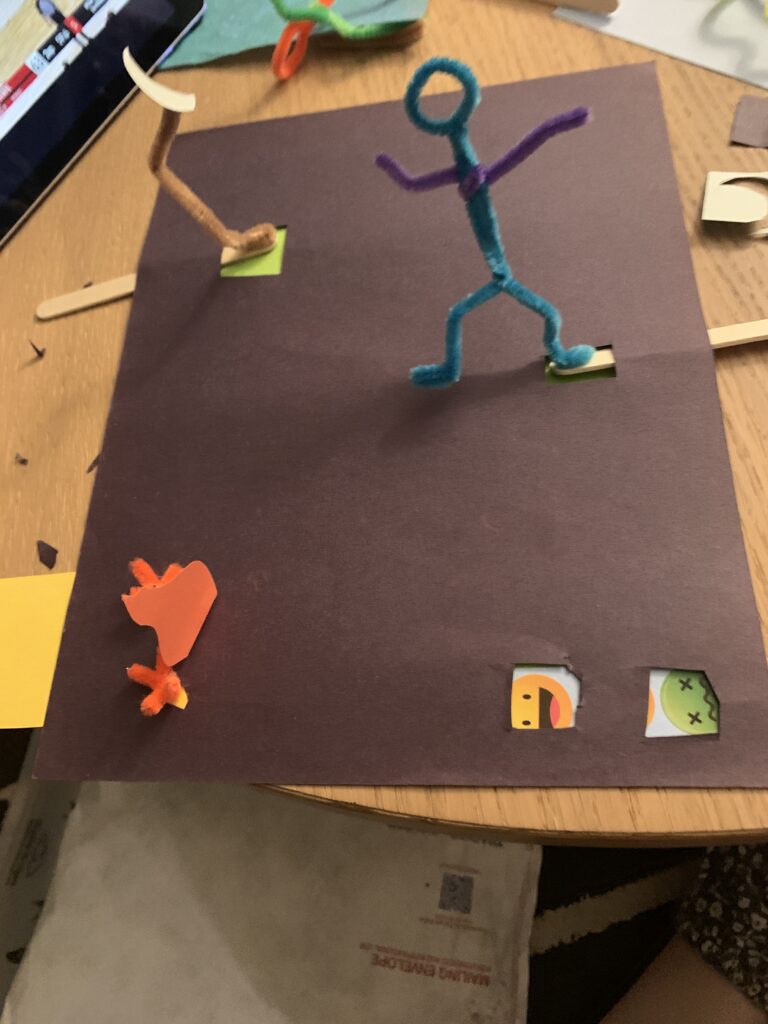 A piece of construction paper with pipe cleaner figures standing on it.