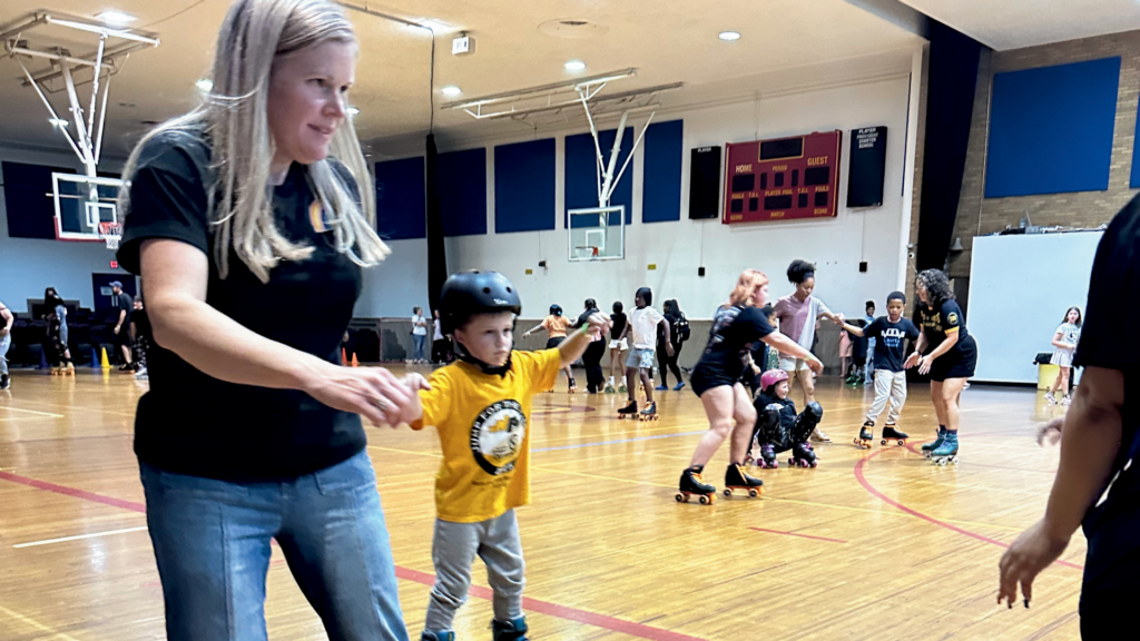 A blond woman and a young boy skate in a school gymnasium with more people skating in the background.