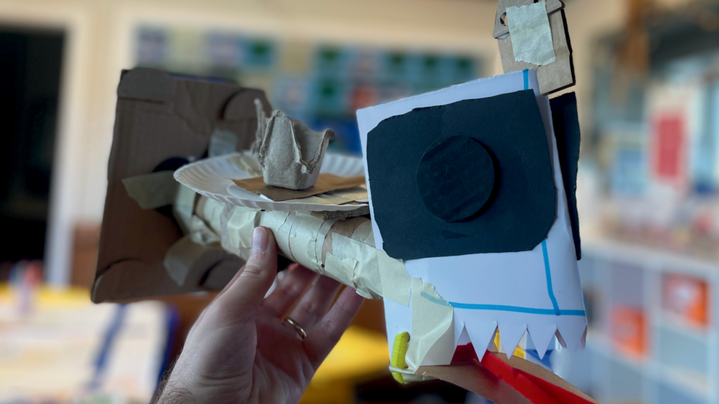 A hand holds up an abstract craft constructed of paper and masking tape.